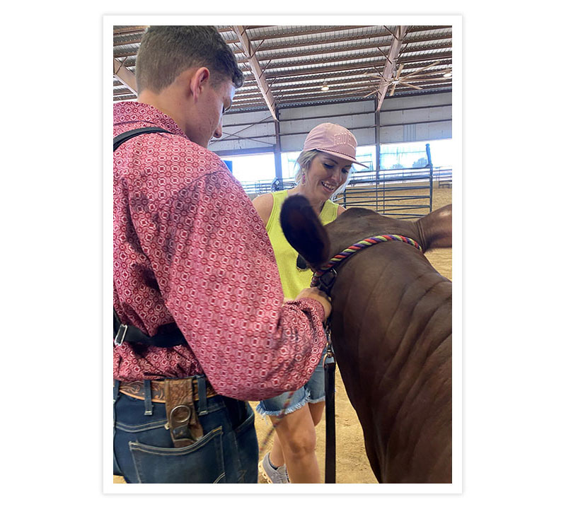 A woman with blond hair wearing a pink ballcap, a bright yellowish green tank top, and jean shorts is helping a student clean up his show cow. 